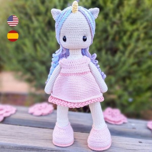 May include: A handmade crochet unicorn doll with a light pink dress, shoes, and a lavender and blue mane. The doll has a cream-colored body, a yellow horn, and black eyes. The doll is approximately 12 inches tall.