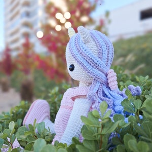 K&ouml;nnte beinhalten: Handgefertigte H&auml;kel-Einhorn-Puppe mit wei&szlig;em K&ouml;rper, lila und blauem Haar und einem rosa Kleid. Die Puppe hat schwarze Augen und sitzt in einem Bett aus gr&uuml;nen Bl&auml;ttern. Der Hintergrund ist verschwommen und zeigt ein Geb&auml;ude und B&auml;ume.