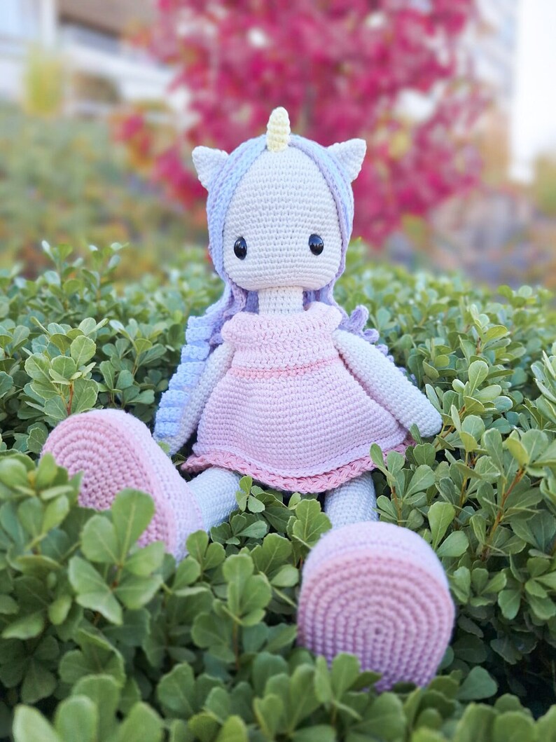 K&ouml;nnte beinhalten: Handgefertigte H&auml;kel-Einhorn-Puppe mit hellblauer und lavendelfarbener M&auml;hne, gelbem Horn und rosa Kleid. Die Puppe sitzt inmitten von gr&uuml;nem Laub, mit einem passenden rosa Fu&szlig; und einem runden Ball.