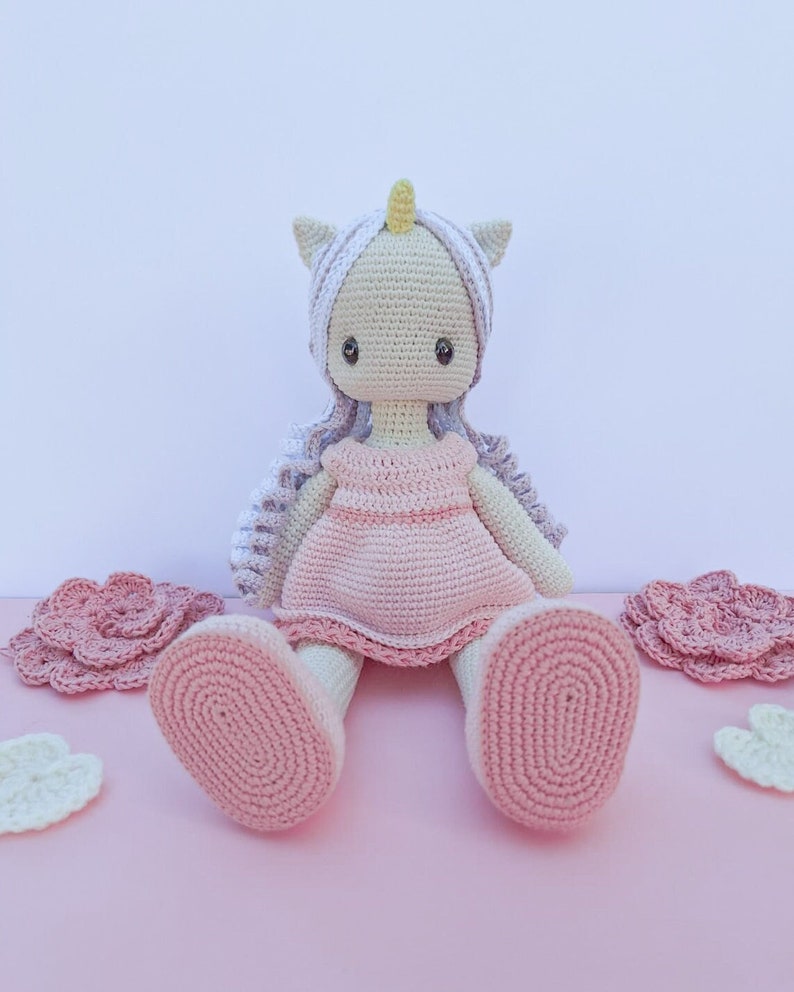 K&ouml;nnte beinhalten: Geh&auml;kelte Einhorn-Puppe mit hellrosa Kleid, wei&szlig;em Haar und gelbem Horn. Die Puppe hat gro&szlig;e, dunkle Augen und sitzt auf einer rosa Oberfl&auml;che. Geh&auml;kelte Blumen und Wolkenformen sind ebenfalls vorhanden.