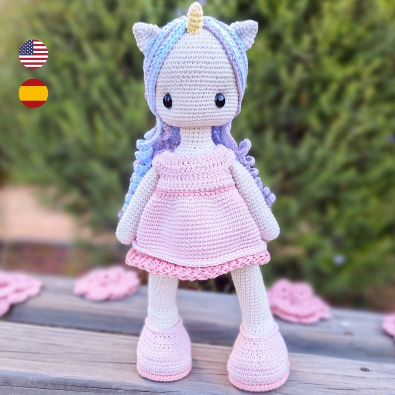 K&ouml;nnte beinhalten: Handgefertigte H&auml;kel-Einhorn-Puppe mit hellrosa Kleid und Schuhen, sowie einer lila-blauen M&auml;hne. Die Puppe hat einen cremefarbenen K&ouml;rper, ein gelbes Horn und schwarze Augen. Die Puppe ist etwa 30 cm gro&szlig;.