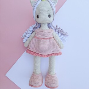 K&ouml;nnte beinhalten: Geh&auml;kelte Einhorn-Puppe mit hellrosa Kleid und passenden Schuhen. Die Puppe hat einen wei&szlig;en K&ouml;rper, graues Haar und ein gelbes Horn. Der Hintergrund ist rosa und wei&szlig;.