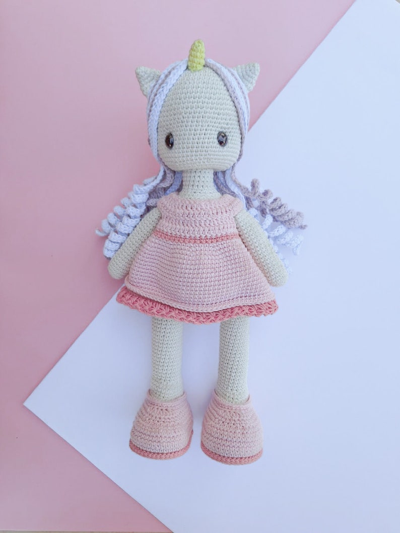 K&ouml;nnte beinhalten: Geh&auml;kelte Einhorn-Puppe mit hellrosa Kleid und passenden Schuhen. Die Puppe hat einen wei&szlig;en K&ouml;rper, graues Haar und ein gelbes Horn. Der Hintergrund ist rosa und wei&szlig;.