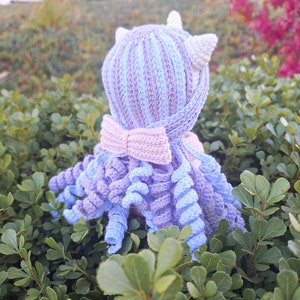 K&ouml;nnte beinhalten: Handgefertigte H&auml;kel-Oktopus-Puppe in Blau-, Lila- und Rosat&ouml;nen. Die Puppe hat einen gestreiften Kopf mit kleinen wei&szlig;en Ohren und einer rosa Schleife. Die Tentakel sind lockig, vor gr&uuml;nem Laub.
