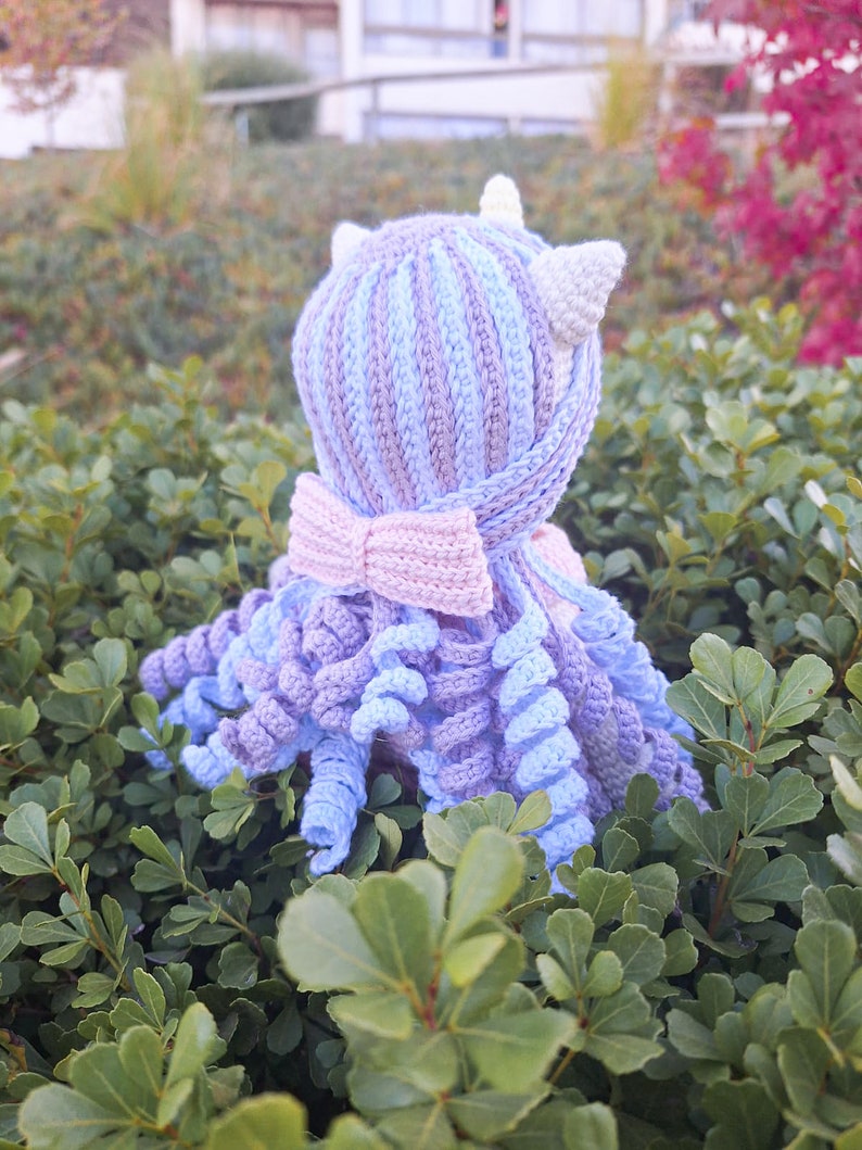 K&ouml;nnte beinhalten: Handgefertigte H&auml;kel-Oktopus-Puppe in Blau-, Lila- und Rosat&ouml;nen. Die Puppe hat einen gestreiften Kopf mit kleinen wei&szlig;en Ohren und einer rosa Schleife. Die Tentakel sind lockig, vor gr&uuml;nem Laub.