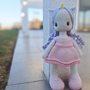 K&ouml;nnte beinhalten: Handgemachte H&auml;kel-Einhorn-Puppe mit hellgrauem Kopf, lavendelfarbenem Haar und gelbem Horn. Die Puppe tr&auml;gt ein rosa Kleid und Schuhe. Die Puppe steht neben einer wei&szlig;en S&auml;ule.