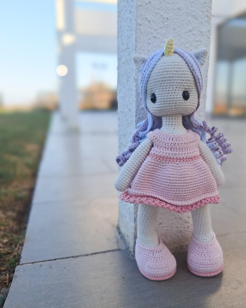 K&ouml;nnte beinhalten: Handgemachte H&auml;kel-Einhorn-Puppe mit hellgrauem Kopf, lavendelfarbenem Haar und gelbem Horn. Die Puppe tr&auml;gt ein rosa Kleid und Schuhe. Die Puppe steht neben einer wei&szlig;en S&auml;ule.