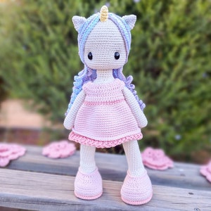 K&ouml;nnte beinhalten: Handgemachte H&auml;kel-Einhorn-Puppe mit hellgrauem Kopf, blau-violettem Haar und gelbem Horn. Die Puppe tr&auml;gt ein rosa Kleid und Schuhe. Die Puppe steht auf einer Holzoberfl&auml;che.