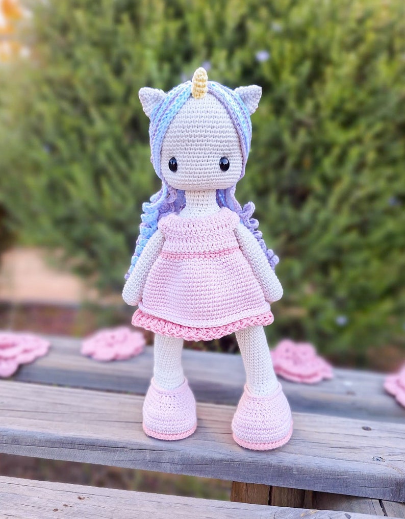 K&ouml;nnte beinhalten: Handgemachte H&auml;kel-Einhorn-Puppe mit hellgrauem Kopf, blau-violettem Haar und gelbem Horn. Die Puppe tr&auml;gt ein rosa Kleid und Schuhe. Die Puppe steht auf einer Holzoberfl&auml;che.