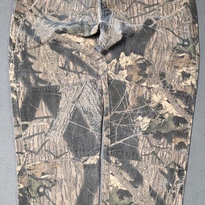 May include: Camouflage-patterned trousers in shades of brown, green, and grey. The trousers feature a classic five-pocket design and a straight leg cut. The fabric has a textured appearance, and the overall style is casual.