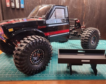 May include: Black and red toy truck with a black bed. The truck has large tires and a black and silver wheel. The truck is sitting on a green cutting mat.