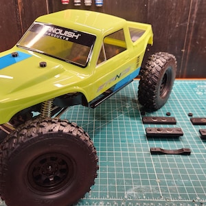 May include: A lime green and black remote control truck with large black tires. The truck has blue accents and the text "VANQUISH PRODUCTS" on the windshield. Several black parts are arranged next to the truck on a green cutting mat.