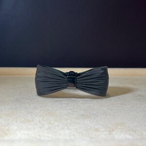 May include: A black bow-shaped hair clip with a textured fabric finish.