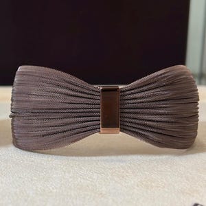 Pode incluir: Um bracelete de metal castanho com um design de laço. O bracelete é feito de uma malha de metal tecida e tem um fecho de ouro rosa.