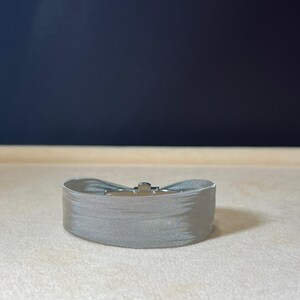 May include: A silver metal belt with a buckle. The belt is made of a woven mesh material.
