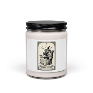 Peut inclure: Une bougie en pot de verre cylindrique avec un couvercle en métal noir, remplie de cire blanc cassé. L'étiquette frontale présente une illustration en noir et blanc style tarot d'une personne en chapeau de sorcière lisant avec un chat, marquée WITCHY ENERGY.