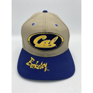 May include: A tan and blue baseball cap featuring the word "Cal" in a navy blue oval with a gold border. The peak is blue, and the word "Berkeley" is embroidered in gold.