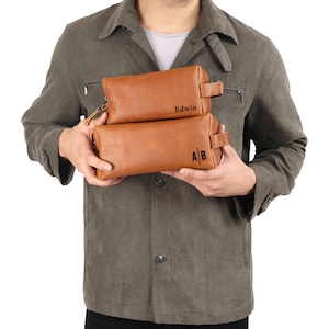 May include: Two brown leather travel bags with zipper closures and handles. The top bag has the name "Edwin" embossed on it. The bottom bag has the initials "A|B" embossed on it.