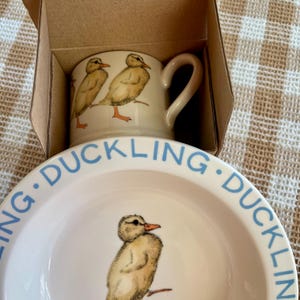 May include: A white ceramic mug and plate set featuring illustrations of yellow ducklings. The mug has two ducklings, and the plate has one. The word "DUCKLING" is written in blue around the plate's rim. The set is in a cardboard box.