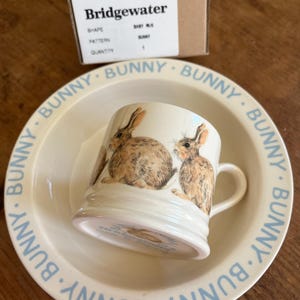 May include: A cream-colored ceramic baby mug with a bunny design, resting in a matching bowl. The bowl and mug feature a light blue "BUNNY" text around the rim. The mug has a handle and is decorated with brown and tan bunnies.