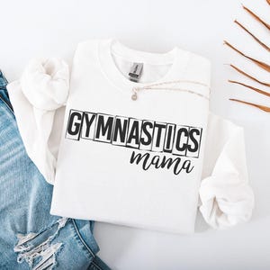 May include: A white crewneck sweatshirt featuring the black text GYMNASTICS in a block-letter style, with the word mama written in a cursive script underneath. The sweatshirt is paired with light blue distressed denim jeans.
