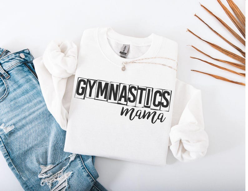 May include: A white crewneck sweatshirt featuring the black text GYMNASTICS in a block-letter style, with the word mama written in a cursive script underneath. The sweatshirt is paired with light blue distressed denim jeans.