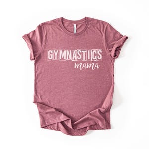 May include: A heathered mauve short-sleeve t-shirt featuring the word GYMNASTICS in a white, blocky, distressed font on the upper chest, with the word mama written below it in a white, cursive, script-style font. The shirt has a crew neckline.