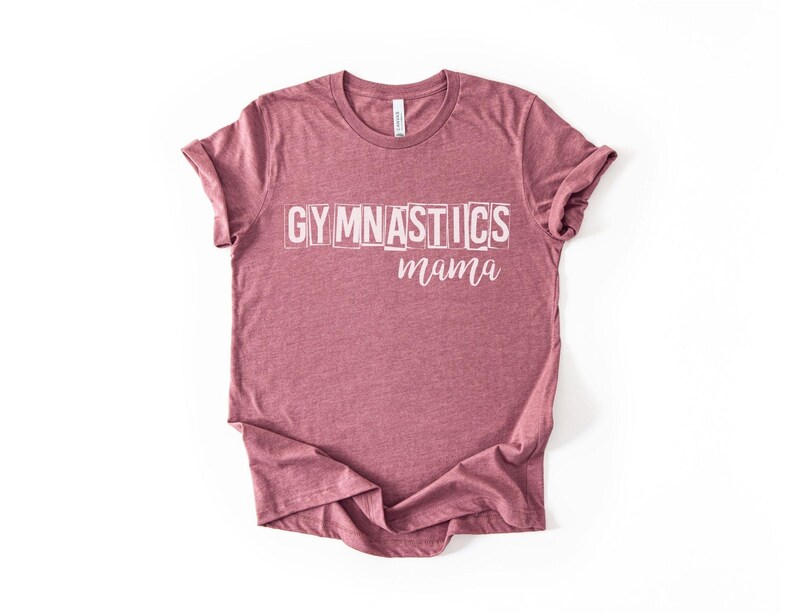 May include: A heathered mauve short-sleeve t-shirt featuring the word GYMNASTICS in a white, blocky, distressed font on the upper chest, with the word mama written below it in a white, cursive, script-style font. The shirt has a crew neckline.
