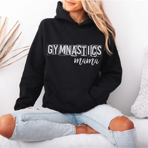 May include: A black hooded sweatshirt featuring a front kangaroo pocket and long sleeves. The chest displays the word GYMNASTICS in white block letters, with the word mama written in a white cursive script font directly underneath the text.