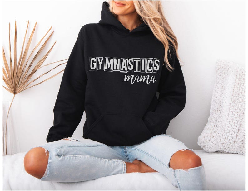 May include: A black hooded sweatshirt featuring a front kangaroo pocket and long sleeves. The chest displays the word GYMNASTICS in white block letters, with the word mama written in a white cursive script font directly underneath the text.