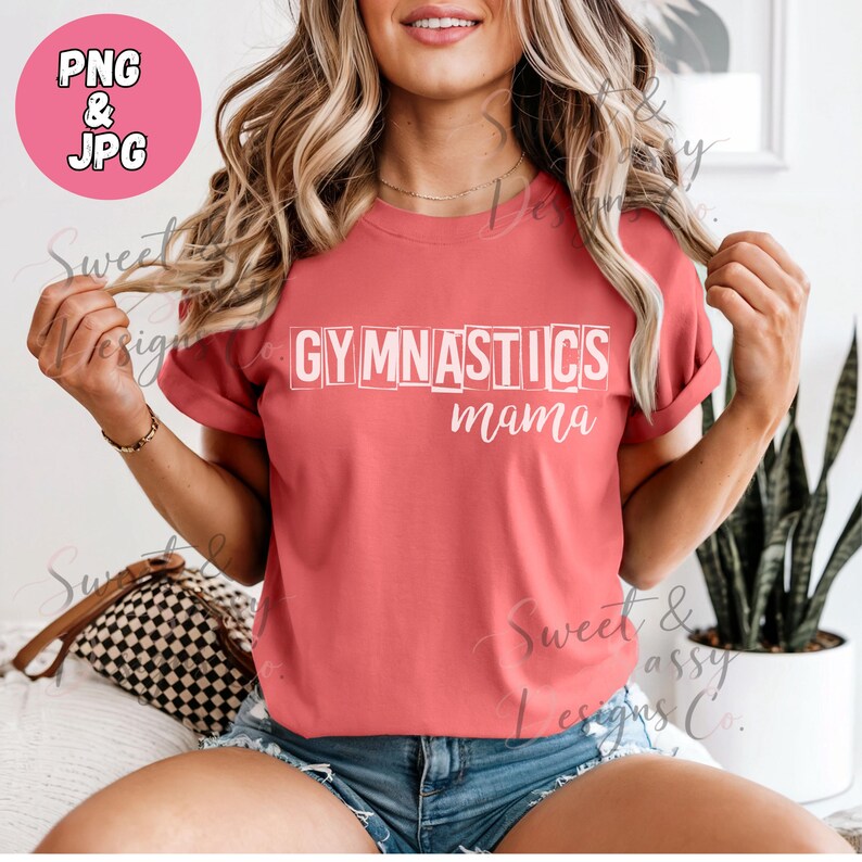 May include: A coral-coloured short-sleeve t-shirt featuring the white text GYMNASTICS in a blocky, outlined font with the word mama written in a cursive script underneath. The shirt has a crew neckline and is worn with blue denim shorts.
