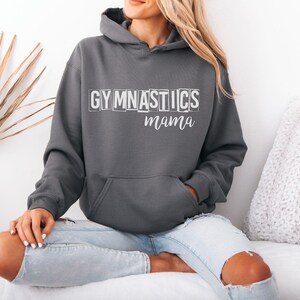 May include: A dark charcoal grey hooded sweatshirt featuring the word GYMNASTICS in a white block-letter font with a distressed effect, followed by the word mama in a white cursive script. The garment includes a front kangaroo pocket and long sleeves.