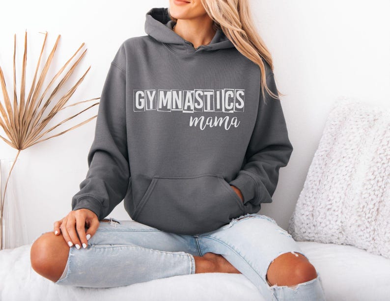 May include: A dark charcoal grey hooded sweatshirt featuring the word GYMNASTICS in a white block-letter font with a distressed effect, followed by the word mama in a white cursive script. The garment includes a front kangaroo pocket and long sleeves.