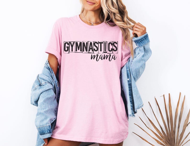 May include: A light pink short-sleeved t-shirt featuring the word GYMNASTICS in bold black block letters with a stacked effect, followed by the word mama in a black cursive script font. The shirt is worn with a light blue denim jacket.