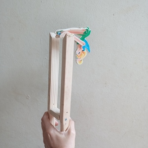 Peut inclure: Un jouet en bois clair avec un personnage de dessin animé attaché. Le personnage a des cheveux blonds, une robe bleue et des ailes vertes. Le jouet est tenu par une main.