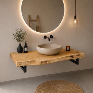 May include: A wall-mounted live edge wooden vanity countertop featuring a round stone vessel sink, a black wall-mounted faucet, a black soap dispenser, a small potted plant, and a candle, all positioned beneath a large round backlit mirror.