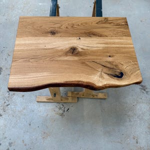Op de afbeelding: Een vierkante houten tafel met een natuurlijke rand. Het tafelblad is gemaakt van lichtbruin hout met zichtbare nerven en knopen. De tafel wordt ondersteund door een houten voet en metalen steunen. De tafel meet ongeveer 61 cm vierkant.