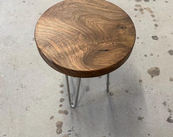 May include: A round wooden side table with a natural wood grain finish. The tabletop is supported by three silver hairpin legs. The table is approximately 18 inches in diameter and 18 inches tall.