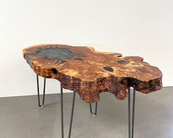 May include: A live edge coffee table with a unique, organic shape. The tabletop is made of natural wood with a dark, epoxy resin-filled section. The table is supported by four metal hairpin legs, creating a modern, rustic aesthetic.