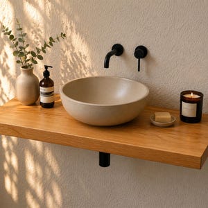 Puede incluir: Un mueble de baño moderno con una estantería flotante de madera clara. Cuenta con un lavabo beige, un grifo negro y una vela encendida. Un pequeño jarrón con vegetación y un jabonera también están en la estantería.