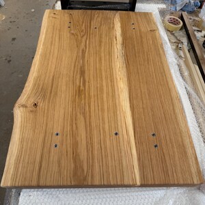 May include: A rectangular light brown wooden tabletop featuring a natural live edge on the left side. The surface displays a prominent wood grain pattern with visible knots and is secured to a black metal frame with six visible silver screws.