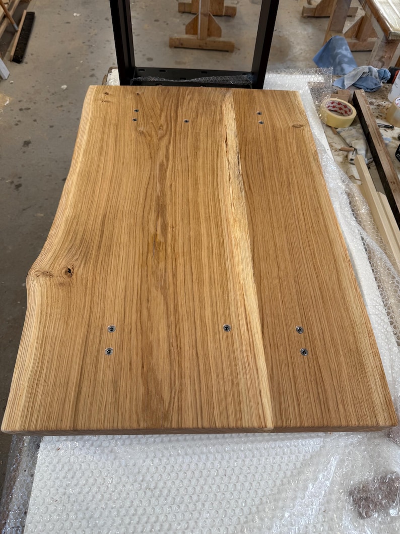 May include: A rectangular light brown wooden tabletop featuring a natural live edge on the left side. The surface displays a prominent wood grain pattern with visible knots and is secured to a black metal frame with six visible silver screws.