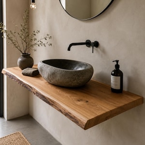 Può includere: Un lavabo in pietra naturale poggia su un ripiano sospeso in legno con bordo grezzo. Un rubinetto a parete nero opaco è posizionato sopra il lavabo. Un dispenser di sapone in vetro scuro e un piccolo vaso in ceramica si trovano sul legno.