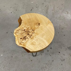Könnte beinhalten: Ein runder Beistelltisch aus Naturholz mit einer Baumkante. Das hellfarbene Holz hat dunkle Flecken und ein einzigartiges Maserungsmuster. Der Tisch wird von drei Metall-Haarnadelbeinen getragen. Der Tisch hat einen Durchmesser von etwa 46 cm.