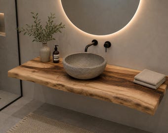 May include: A live edge wooden vanity countertop featuring a round stone vessel sink, a matte black wall-mounted faucet, a ceramic vase with green foliage, a dark brown soap dispenser, and a stack of folded beige towels under a round mirror.