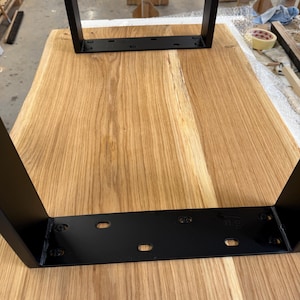 May include: A rectangular light brown wooden tabletop featuring a natural wood grain pattern. Two black metal U-shaped legs are attached to the underside of the wood surface with black metal mounting plates secured by visible screws.