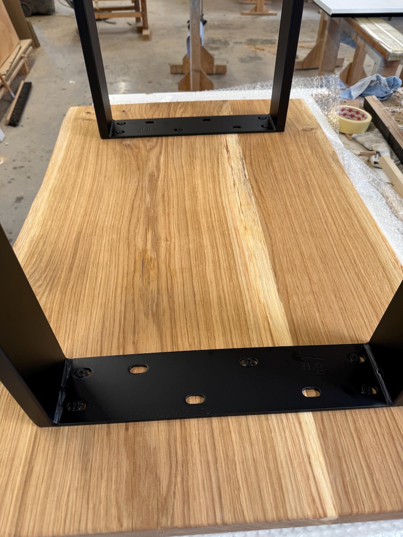 May include: A rectangular light brown wooden tabletop featuring a natural wood grain pattern. Two black metal U-shaped legs are attached to the underside of the wood surface with black metal mounting plates secured by visible screws.