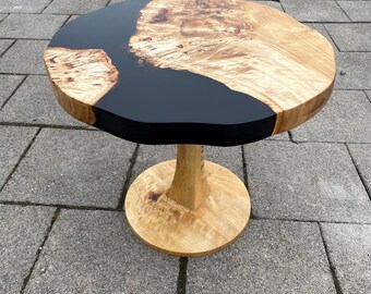 May include: A round wooden side table with a natural edge and a black epoxy resin river. The table is supported by a pedestal base. The wood has a light brown color with a unique grain pattern.