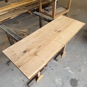 Puede incluir: Una mesa de madera clara sin terminar con un borde natural. La tapa rectangular está soportada por una base de madera sencilla. Otra mesa similar es visible en el fondo.