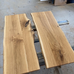 May include: Two rectangular light brown wooden boards with natural live edges. The wood features visible grain patterns, knots, and dark organic markings. The surfaces are smooth and flat, showcasing the raw texture and light tan color of the timber.